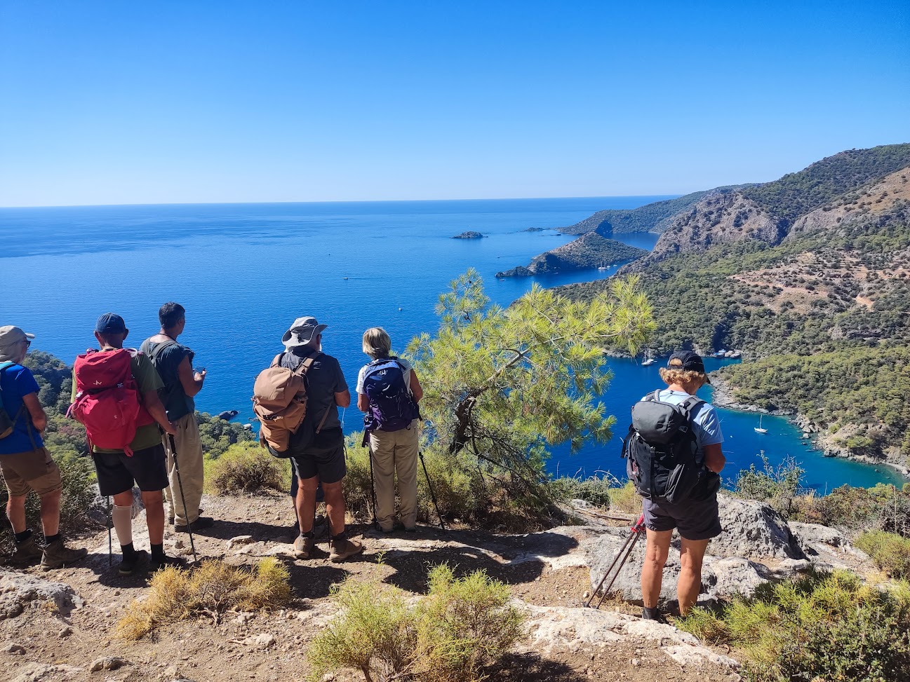 Groupe de randonneurs avec sacs à dos regardant une baie bleue depuis une crête rocheuse bordée de collines boisées
