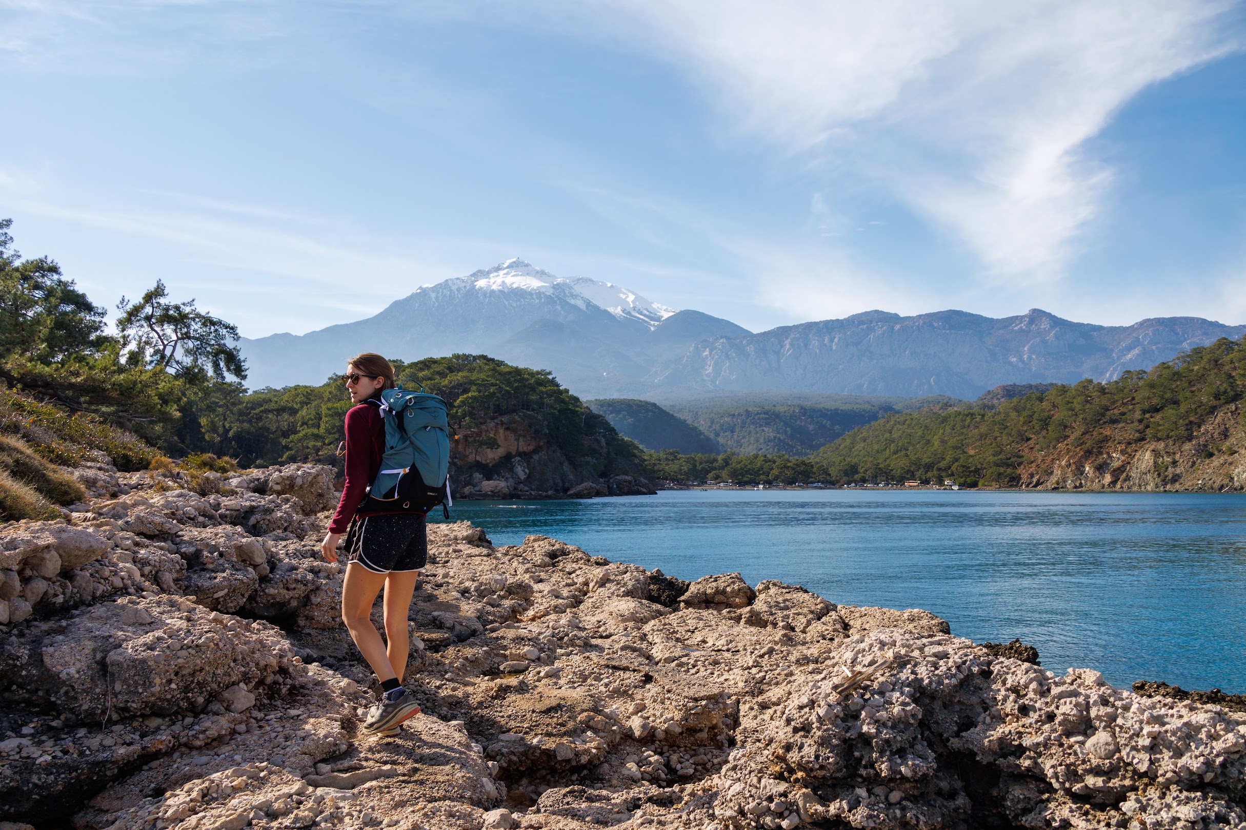 A hiker on a rocky limestone headland above a bay, with snow-capped mountains behind