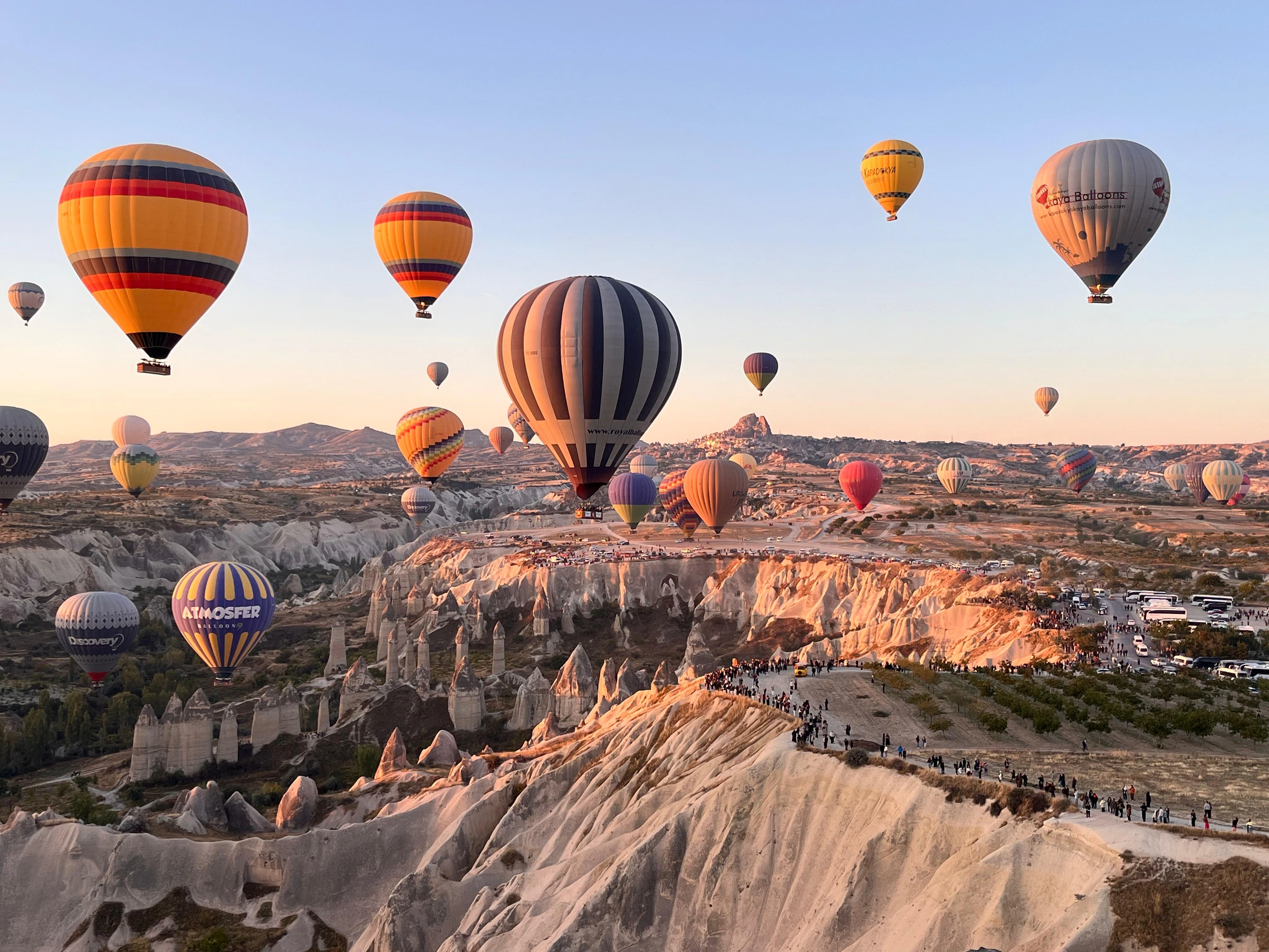 De nombreuses montgolfières colorées survolent un paysage de roches érodées et de cheminées de fée avec des groupes de personnes au sol.