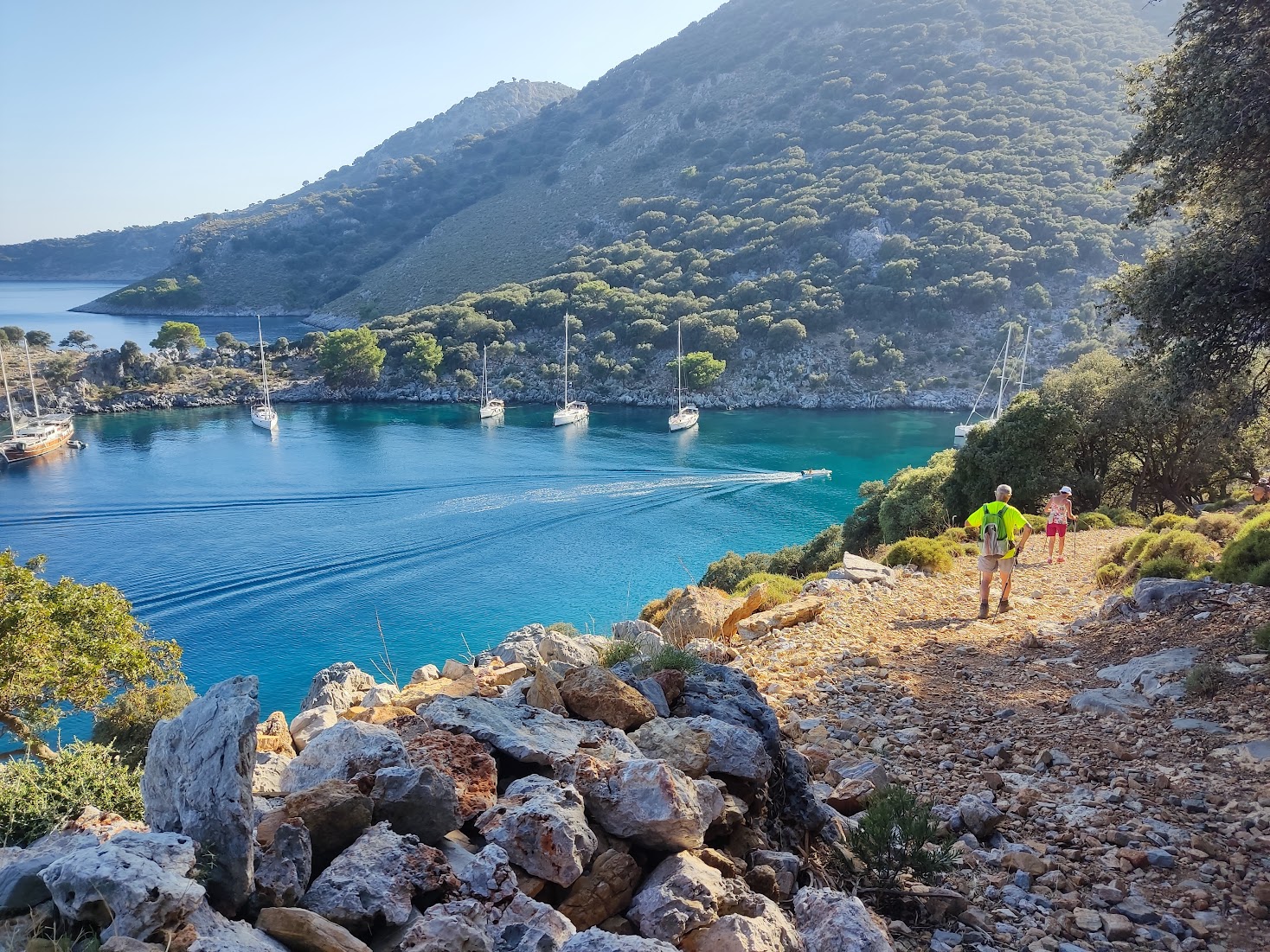 Deux personnes marchent sur un sentier rocheux au-dessus d'une baie turquoise où des voiliers sont ancrés, entourée de collines boisées.