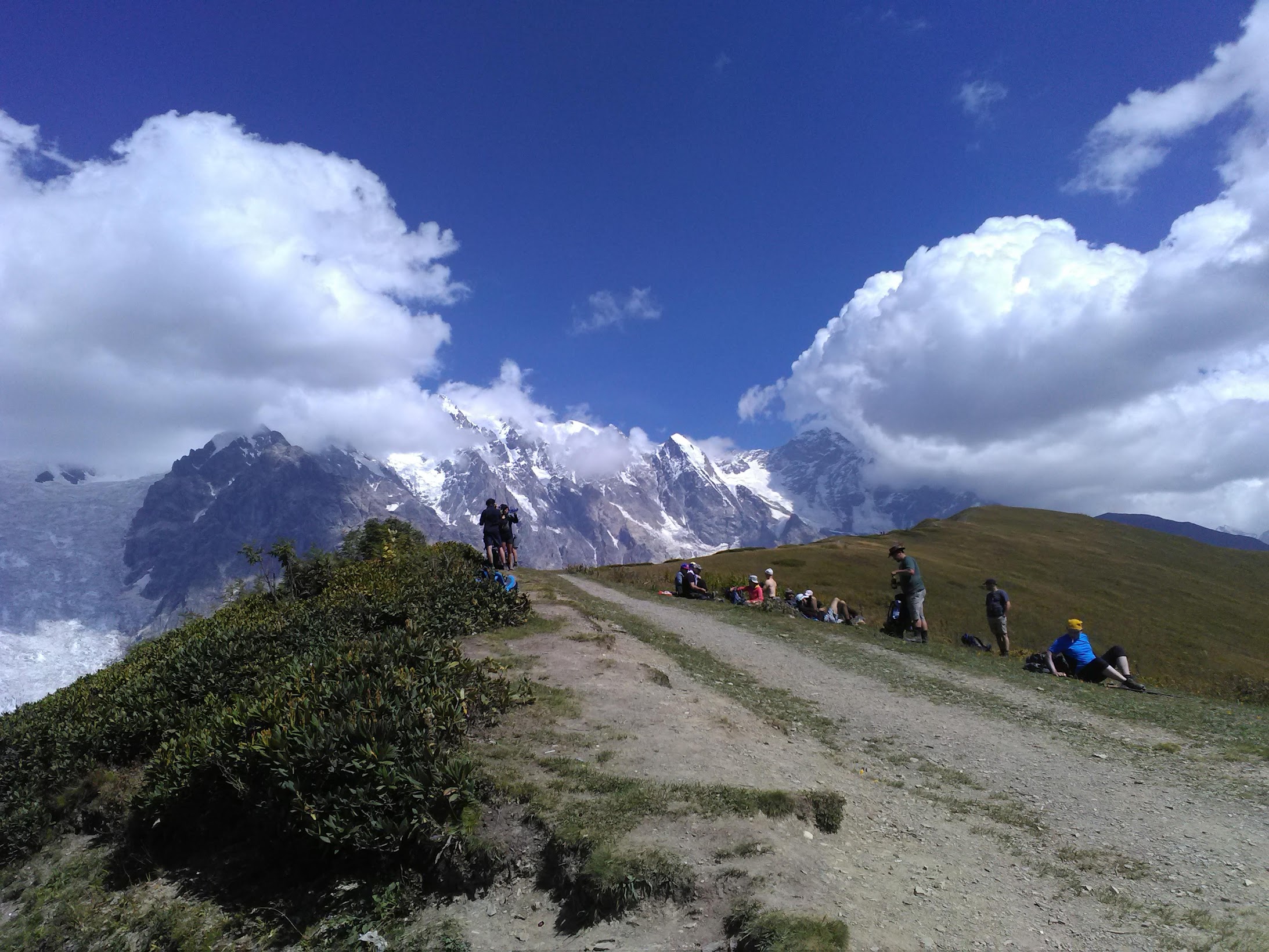 Groupe de randonneurs sur un sentier herbeux face à des sommets enneigés partiellement couverts de nuages, sous un ciel bleu.