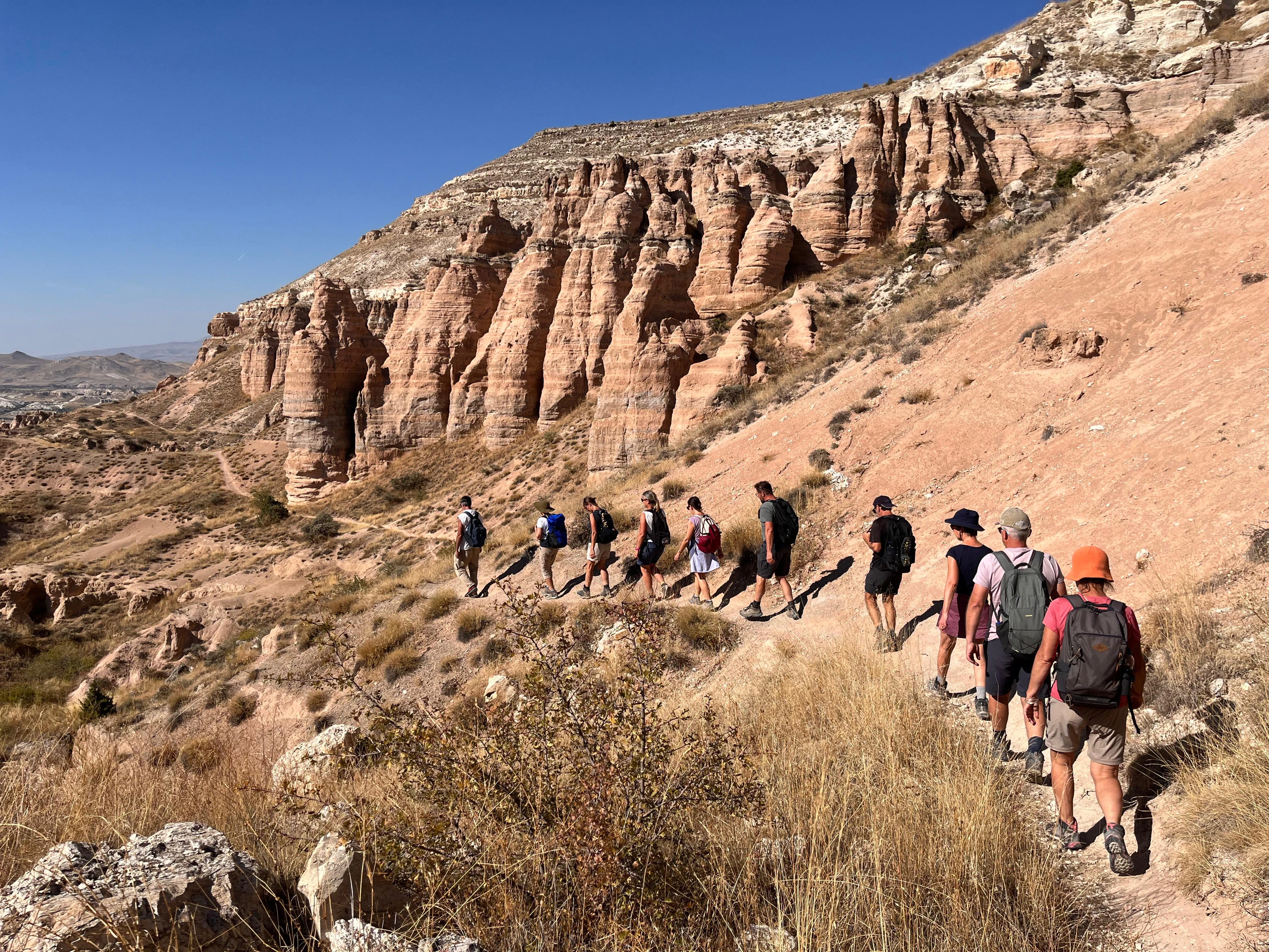 Groupe de randonneurs avec sacs à dos marchant en file sur un sentier poussiéreux près de falaises rocheuses orangées sous un ciel bleu.