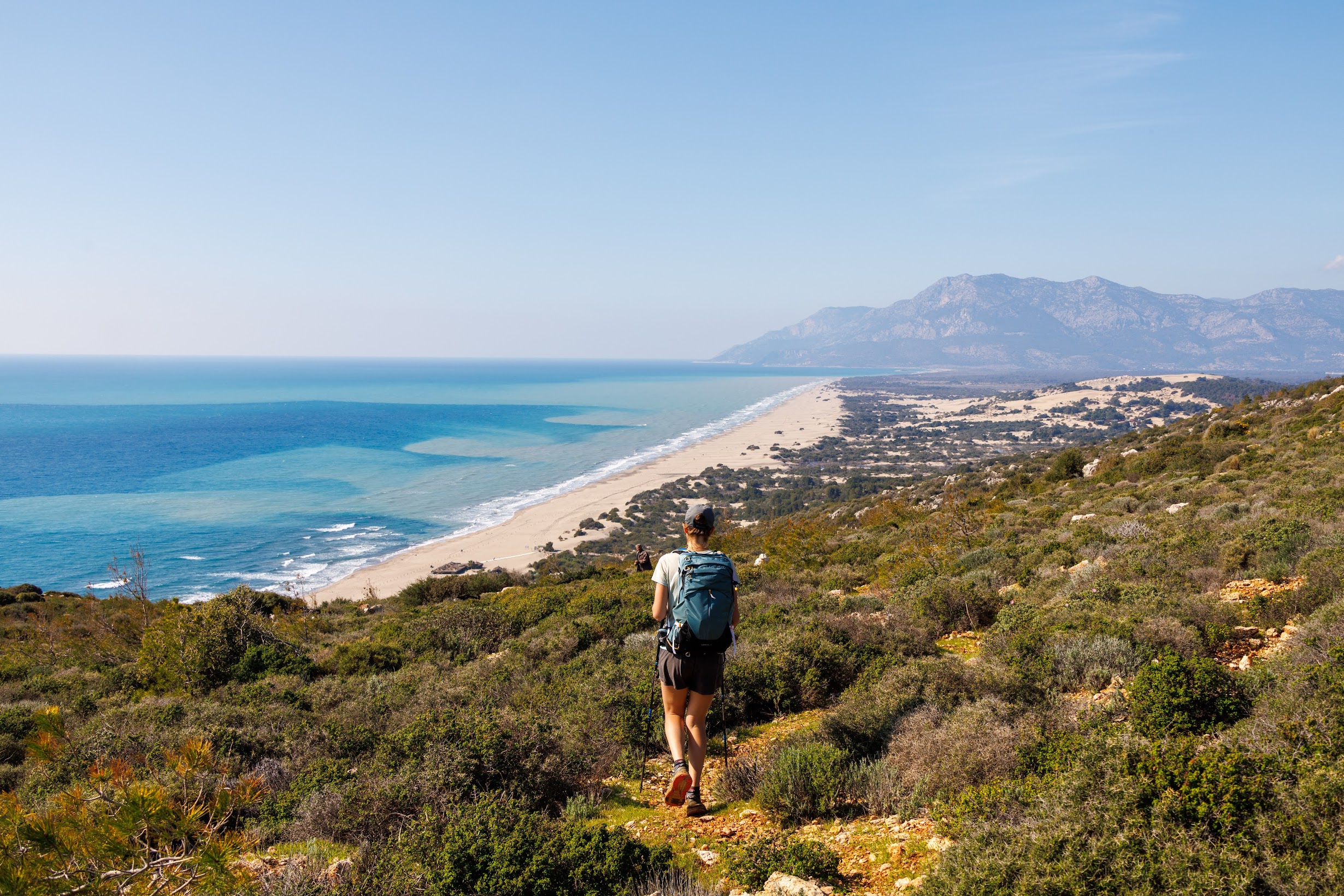 A walker with a backpack on a scrub-covered path above the long sandy beach of Patara