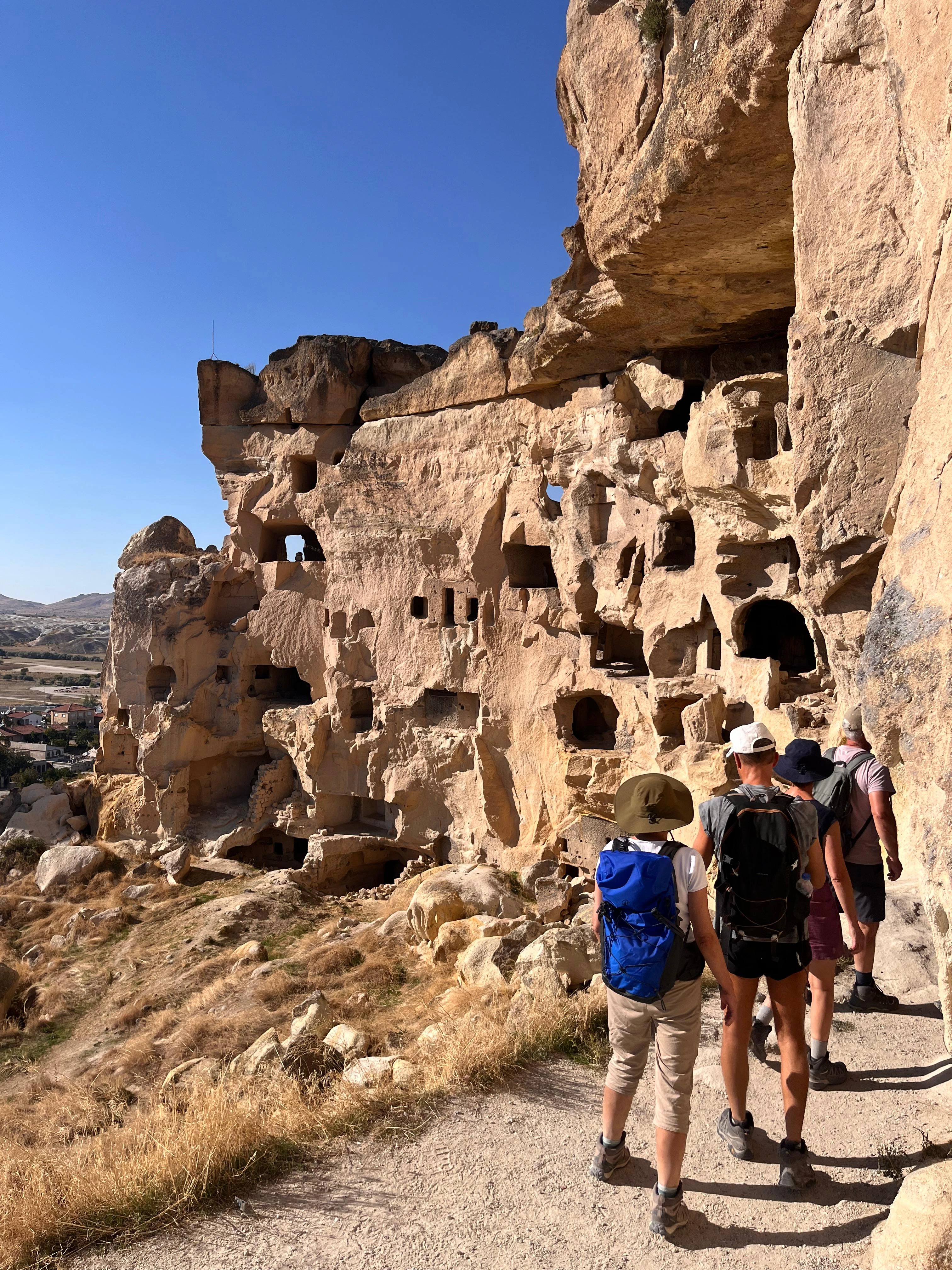 Groupe de randonneurs avec sacs à dos marchant sur un sentier le long d'une falaise creusée d'habitations rupestres sous un ciel bleu.
